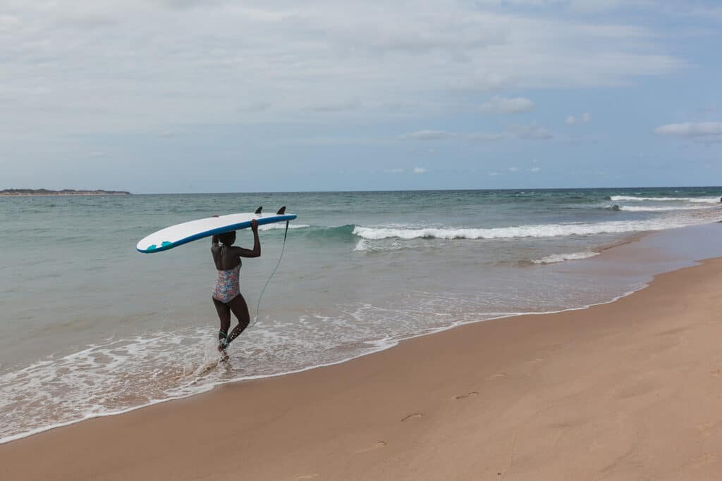 Beautiful young African surfer girl walking with surfboard to th
